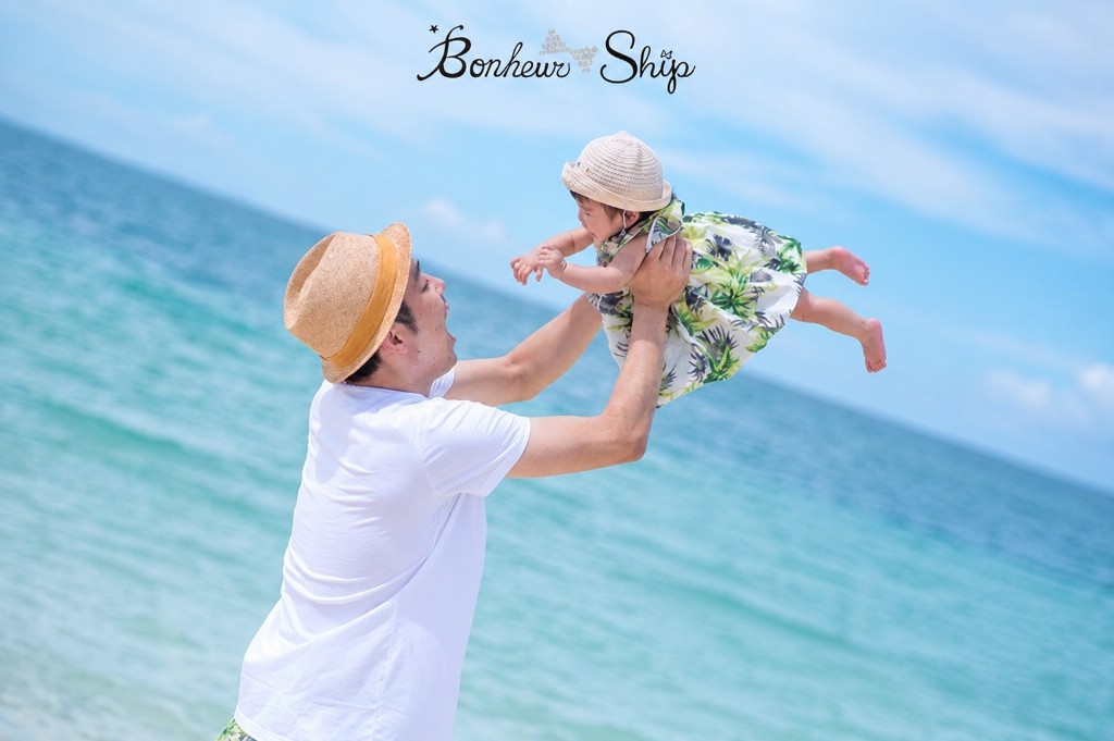 海辺で赤ちゃんを抱き上げて笑顔を見せる親子。Bonheur Ship